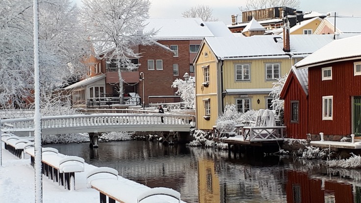 Snötäckta bänkar invid å, i bakgrunden äldre trähus.