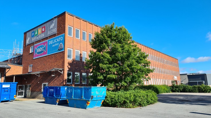 Tegelbyggnad i tre våningar. Blå himmel. I förgrunden står containrar.