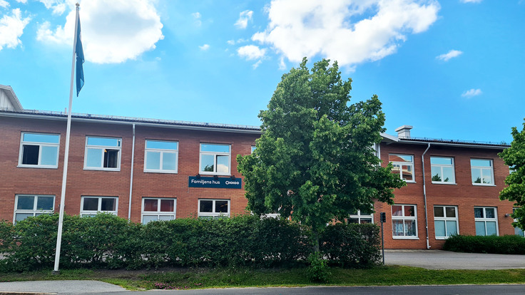 En röd tegebyggnad med texten Familjens hus. Blå himmel, träd och häck i förgrunden.