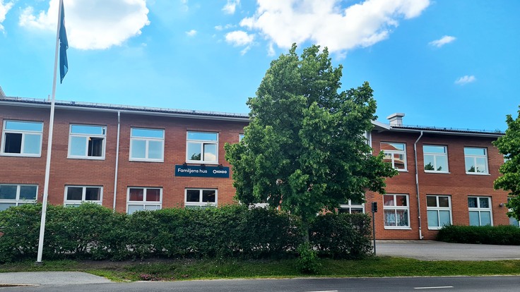 En röd tegebyggnad med texten Familjens hus. Blå himmel, träd och häck i förgrunden.