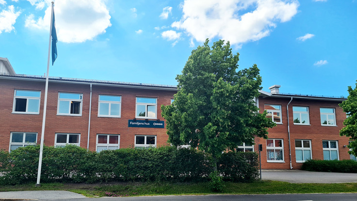 En röd tegebyggnad med texten Familjens hus. Blå himmel, träd och häck i förgrunden.