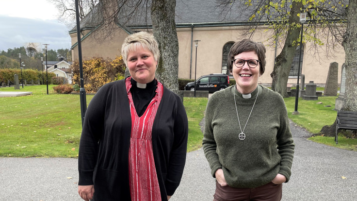 Två leende kvinnor står framför kyrka med kyrkogård.