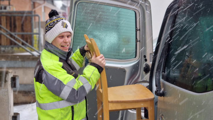 Brukare inom daglig verksamhet, klädd i reflexjacka lastar in en möbel i bageluckan på en bil. Snön yr runt honom.