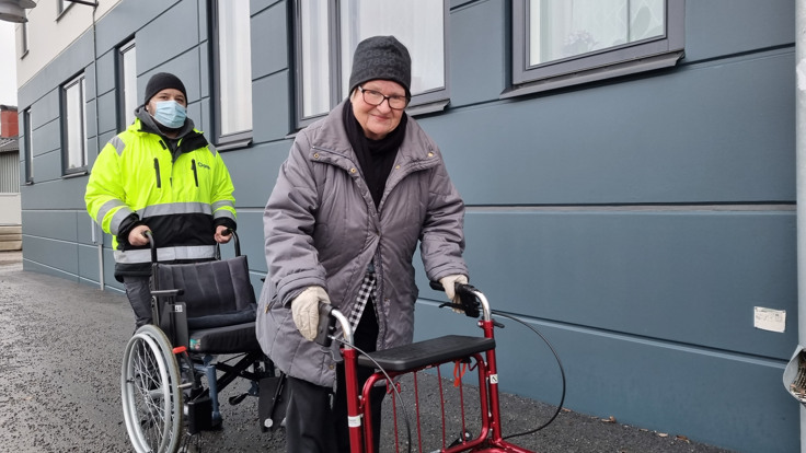 Man i Tiohundraprofilerad jacka med rullstol på promenad med en äldre kvinna i grå jacka och rullator.