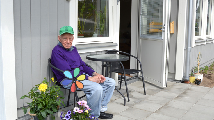 Man sitter på stol framför grått hus. Blomkrukor i förgrunden. 