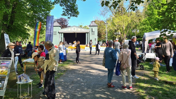 Människor går omkring eller står och pratar med varandra i en park. I bakgrunden en scen. Det är vackert väder.