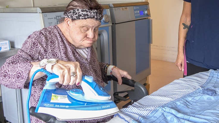 Boende som sitter och stryker. Har ett stort strykjärn i handen.